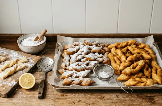 Bugnes lyonnaises maison tout juste frites sur une plaque, moitié saupoudrées de sucre glace, pâte nouée sur planche