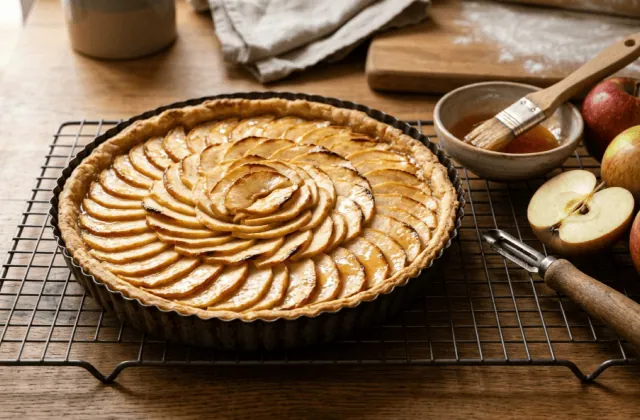 Tarte aux pommes maison en rosace dorée et nappée sur une grille, pinceau, pommes entières et coupées