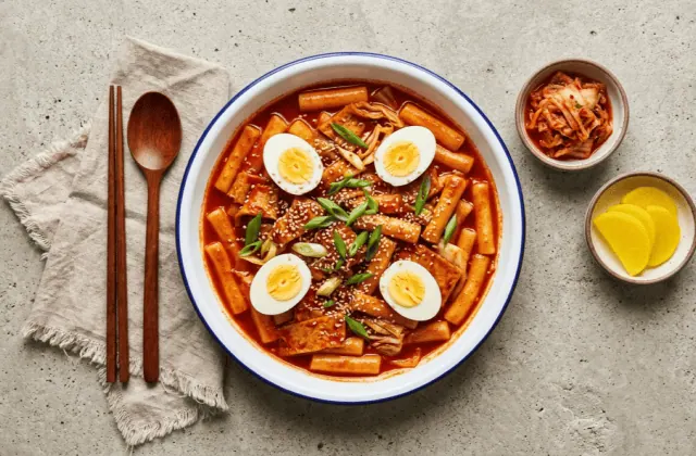 Tteokbokki épicé maison avec gâteaux de poisson, œufs durs, cébette et sésame, servi avec kimchi et radis jaune